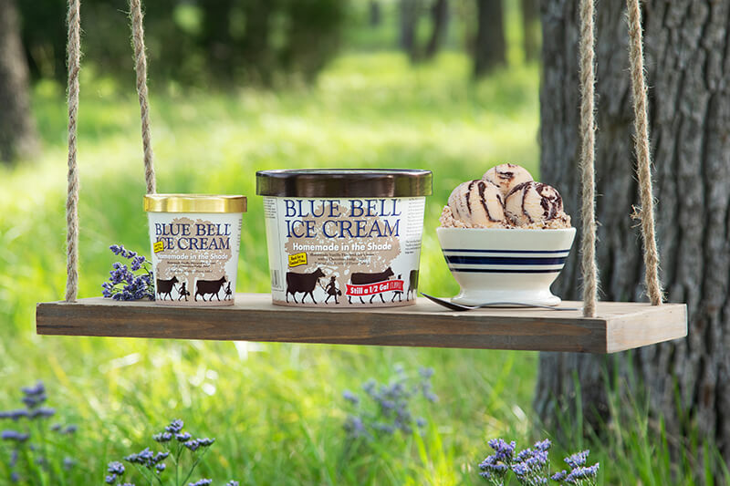 Blue Bell Ice Cream pints and a bowl of scoops on a wooden swing under Brenham oaks
